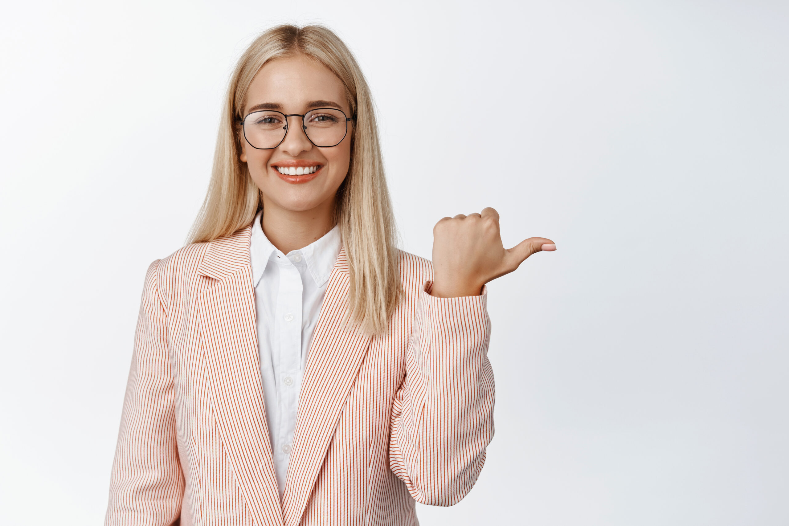 Young saleswoman in glasses and suit, pointing finger right, smiling and showing advertisement, white background.